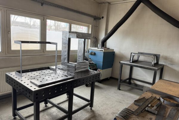 Ein rohes Stahlgehäuse wird auf einem professionellen Schweißtisch in der AMBIO Werkstatt in Dorfen bearbeitet. Im Hintergrund ist eine Absauganlage sichtbar.