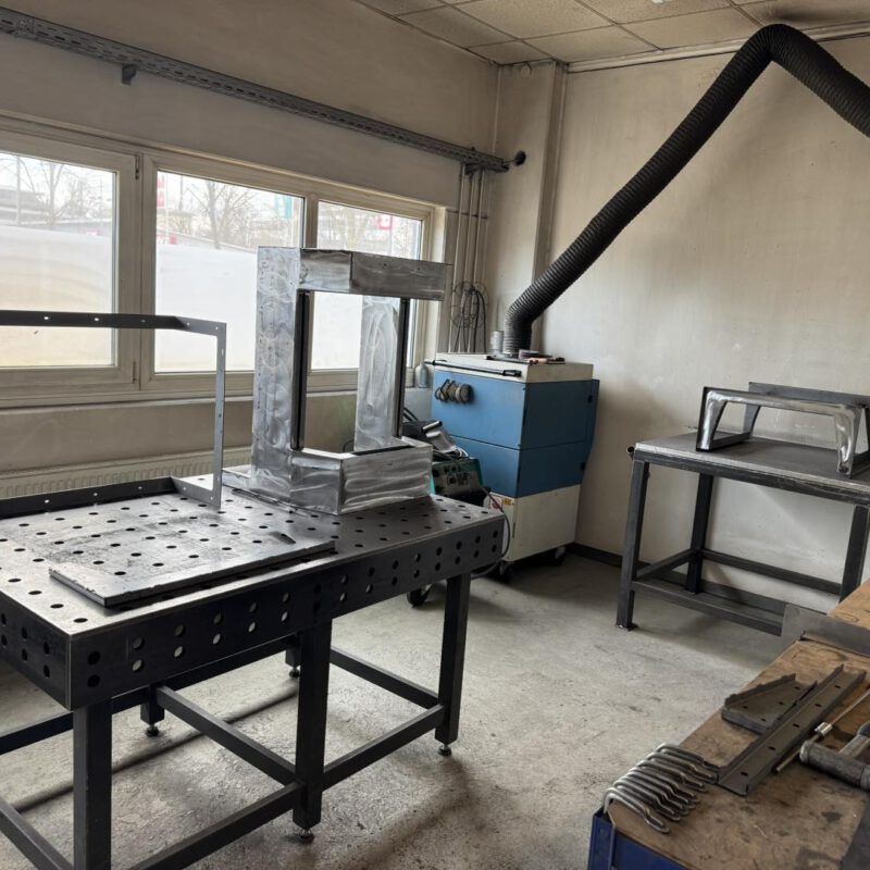 Ein rohes Stahlgehäuse wird auf einem professionellen Schweißtisch in der AMBIO Werkstatt in Dorfen bearbeitet. Im Hintergrund ist eine Absauganlage sichtbar.